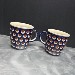 Puede incluir: Dos tazas de cerámica con fondo azul oscuro y un patrón de círculos marrones y blancos. Las tazas tienen un interior blanco y un asa azul. Las tazas son probablemente para café o té.