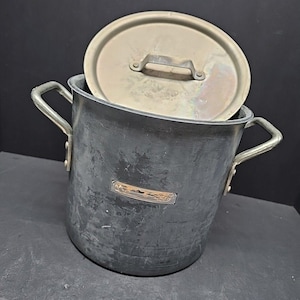 May include: A large, dark gray cooking pot with a matching lid. The pot has two silver handles and a small, rectangular metal plate on the side. The lid is slightly ajar, revealing the interior of the pot.