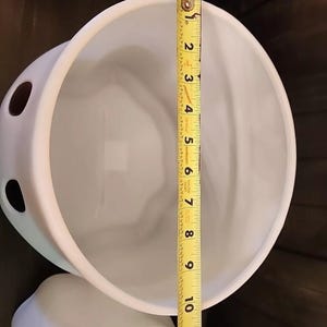 Puede incluir: Un cuenco de cerámica blanca con dos pequeños agujeros en el lateral. El cuenco se mide con una cinta métrica amarilla, mostrando una altura de aproximadamente 25 cm.