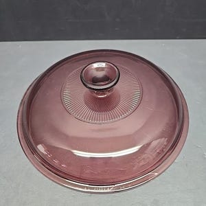 Könnte beinhalten: Ein runder, violetter Pyrex-Deckel mit einem zentralen Knopf. Der Deckel hat eine Reihe konzentrischer Rillen, die vom Knopf ausstrahlen. Das Glas ist durchscheinend und für eine Pyrex-Schüssel.