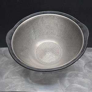 May include: A stainless steel colander with a black rubber rim and handles. The colander has many small holes for draining liquids. The design is simple and functional, suitable for kitchen use.