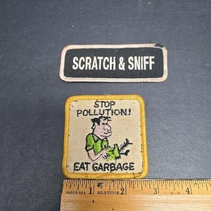 Op de afbeelding: Twee vintage patches. De bovenste patch is zwart met witte tekst die luidt "SCRATCH & SNIFF". De onderste patch heeft een cartoonafbeelding van een man met de tekst "STOP POLLUTION! EAT GARBAGE". Onderaan staat een liniaal.