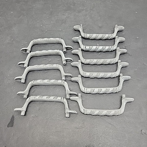 May include: A collection of ten light gray cabinet handles. The handles have a curved design with decorative detailing in the center. The handles are made of metal and are arranged on a gray surface.