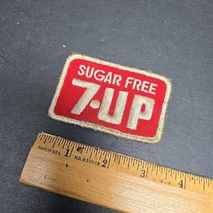 May include: A rectangular red and white embroidered patch with the words "SUGAR FREE" above "7-UP". A wooden ruler with inch markings is next to the patch.