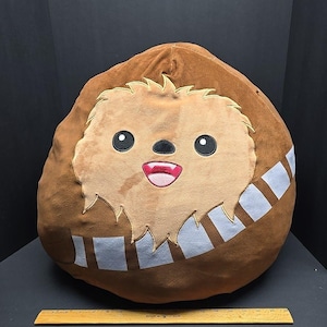 Op de afbeelding: Een bruin pluche kussen in de vorm van een personage uit Star Wars. Het kussen heeft een lichtbruin gezicht met zwarte ogen, een rode mond en een bandolier-ontwerp. Het kussen heeft een diameter van ongeveer 46 cm.