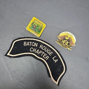 May include: A collection of vintage pins and a patch. The patch is black with white text that reads "Baton Rouge LA Chapter." There is a yellow pin with a Louisiana scene and another pin with an eagle design.