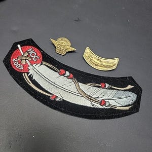 Può includere: Una collezione di toppe e spille Harley-Davidson. La toppa principale presenta una piuma bianca con dettagli rossi e marroni, e il testo "HOG 2005". Sono presenti anche due spille color oro, una con ali e le lettere "H.C."