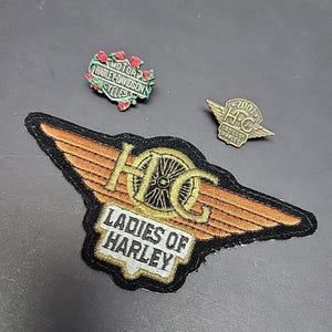 Op de afbeelding: Drie Harley-Davidson patches en pins. Eén patch heeft de tekst "Ladies of Harley" met een wiel- en vleugelontwerp. Een pin heeft de tekst "2007 HFC Ladies of Harley". Een andere pin heeft de tekst "Motor Harley-Davidson Cycles".