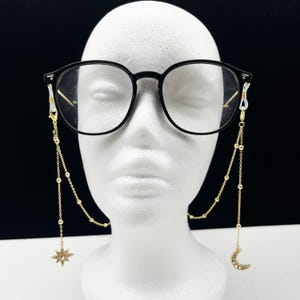 Puede incluir: Cadena de gafas de oro con un dije de estrella y media luna. La cadena está hecha de pequeñas cuentas doradas y tiene un diseño simple.