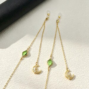 Peut inclure: Une paire de chaînes de lunettes dorées composées de maillons métalliques délicats, ornées chacune d'une petite perle verte à facettes en forme de losange et d'un pendentif croissant de lune serti de minuscules cristaux transparents.
