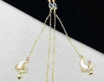 May include: Gold-colored eyeglass chain with white crescent moon and star charms. The chain has small gold beads and connects to the glasses with clear rubber loops. The moon charms have gold accents and a small star charm.