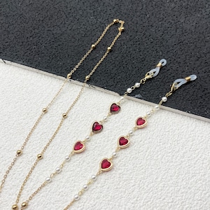 May include: Three gold chains with red heart charms. Two chains feature white pearl beads. The chains have clear plastic clasps for attaching to glasses.