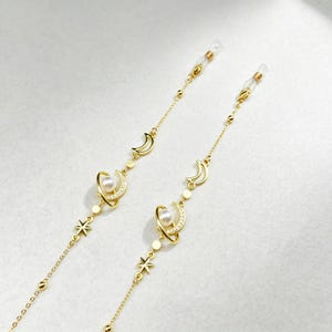 Peut inclure: Une chaîne de lunettes dorée composée de maillons délicats, agrémentée de petites étoiles, de croissants de lune et de pendentifs en forme de planète. Chaque planète comporte une perle blanche entourée d'un anneau de cristaux clairs.