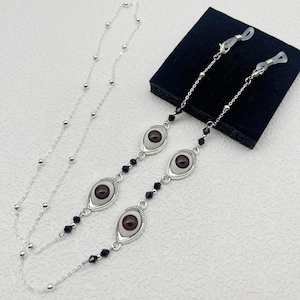 May include: Silver eyeglass chain featuring three eye-shaped charms with brown irises and black accents. The chain has a delicate design with small silver beads and black faceted beads. The eyeglass chain is designed to hold eyeglasses.