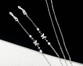May include: Silver-toned eyeglass chains with butterfly and gemstone accents. The chains feature delicate links and are designed to attach to eyeglasses. The butterflies and gemstones add a touch of elegance to the accessory. The background is black.
