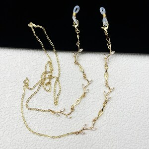 以下が含まれることがあります： 繊細な葉とクリスタルのデザインが施された、ゴールドカラーのメガネチェーン。チェーンには、小さな輝くクリスタルと、メガネに取り付けるための細いチェーンが使用されています。チェーンは首に掛けて着用するように設計されています。