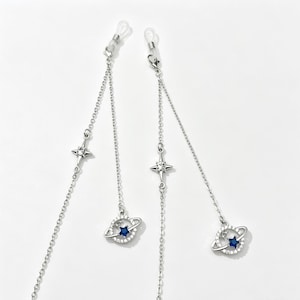 Peut inclure: Une paire de chaînes de lunettes argentées composées de maillons métalliques délicats, chacune ornée d'une breloque étoile à quatre branches avec une pierre claire et d'un pendentif planète circulaire avec une pierre centrale bleu foncé.