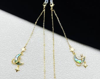 May include: Gold-colored eyeglass chain with a delicate chain design. Each end has a loop to attach to glasses. The chain features a crescent moon pendant with abalone shell inlay and star charms. The chain is approximately 28 inches long.
