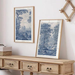 May include: Two framed blue and white landscape prints with trees and a bridge are displayed on a light wood console table. The prints have a classic, vintage aesthetic. The table has drawers with dark metal pulls.