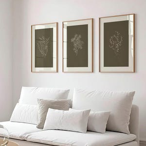 Peut inclure: Trois impressions botaniques encadrées avec un design minimaliste. Chaque impression présente un dessin au trait blanc d'une plante sur un fond vert olive foncé, dans un cadre fin brun clair. Les impressions sont affichées au-dessus d'un canapé blanc avec plusieurs coussins.