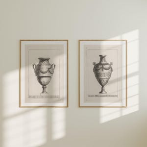 Op de afbeelding: Twee ingelijste prenten met gedetailleerde zwart-wit illustraties van klassieke urnen. De urnen hebben sierlijke handvatten en decoratieve patronen. De prenten zitten in houten frames, wat een vintage esthetiek creëert.