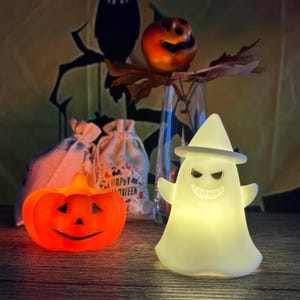 May include: A Halloween display with a glowing orange pumpkin and a white ghost figure wearing a hat. An orange pumpkin with a carved face sits on a vase. Two small bags with "Happy Halloween" text are also present.