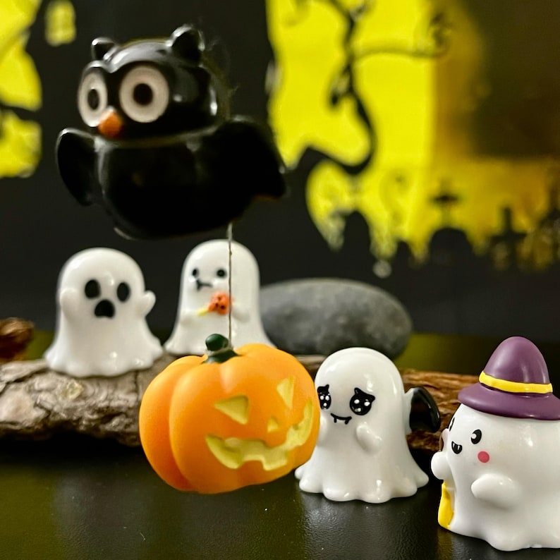 May include: A collection of Halloween-themed figurines, including white ghosts, an orange jack-o'-lantern, and a black owl. The figurines are arranged on a wooden surface against a dark background with yellow accents, creating a festive display.