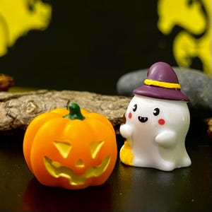 May include: A Halloween-themed image featuring a carved orange pumpkin and a white ghost figurine wearing a purple hat. The pumpkin has a green stem and a carved face. The ghost has a friendly expression and holds a small yellow bag.