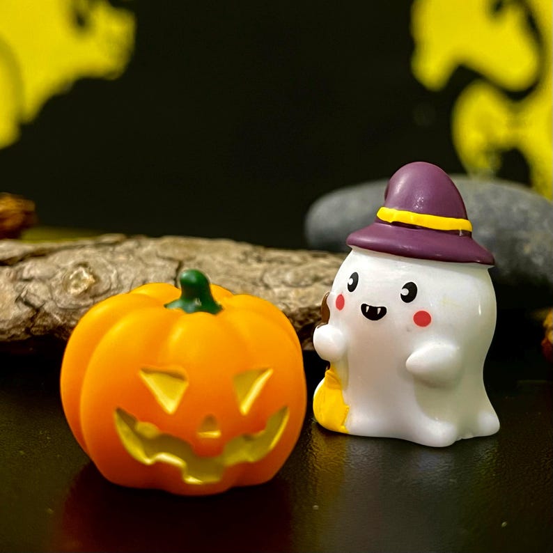 May include: A Halloween-themed image featuring a carved orange pumpkin and a white ghost figurine wearing a purple hat. The pumpkin has a green stem and a carved face. The ghost has a friendly expression and holds a small yellow bag.
