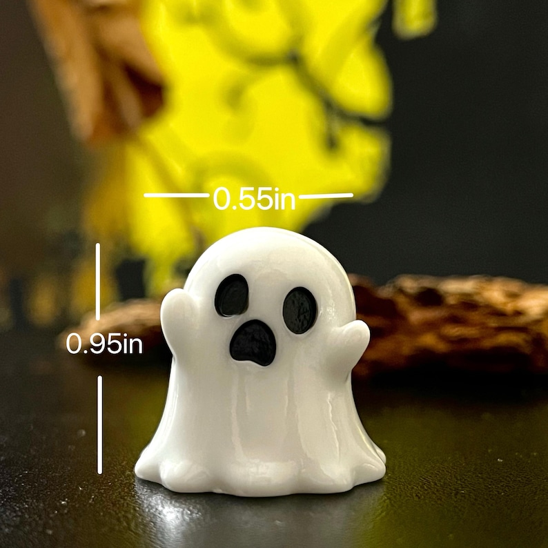 May include: A small, white ghost figurine with black eyes and mouth. The ghost is 0.95 inches tall and 0.55 inches wide. The figurine has a smooth, glossy finish and is likely made of ceramic or resin. The ghost is a Halloween-themed decoration.
