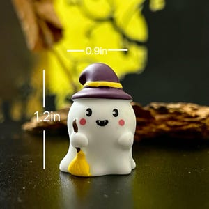 May include: A white ghost figurine, 1.2 inches tall and 0.9 inches wide, wearing a purple witch hat with a yellow band. The ghost has rosy cheeks, a black smiling mouth, and holds a yellow broom. A Halloween-themed decoration.