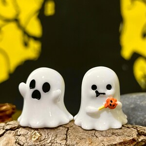 May include: Two small, white ghost figurines with black eyes and mouths. One ghost has an open mouth, and the other holds a small orange and yellow lollipop. The figurines are on a piece of wood.