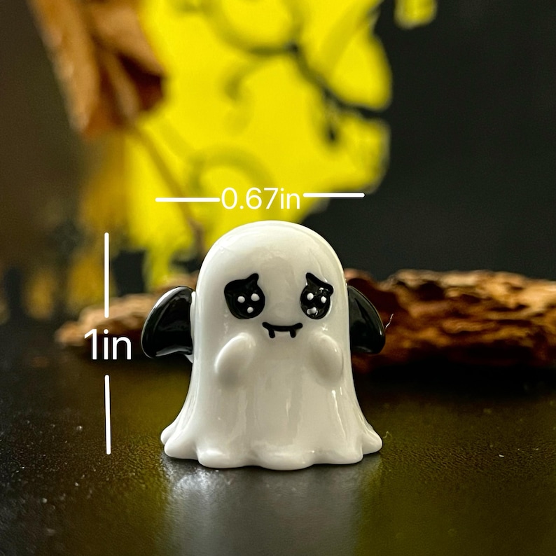 May include: A small, white ghost figurine with black bat-like wings. The ghost has a smiling mouth and two large, round eyes. It measures 1 inch tall and 0.67 inches wide. A Halloween-themed decorative item.