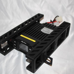 May include: A black AnyTone mobile radio transceiver mounted within a black protective metal cage frame. The device features a front display screen, labeled buttons P1 through P4, a yellow warning sticker on top, and rear power cables.