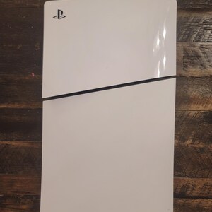 May include: A white gaming console with a black horizontal accent line. The console features a glossy finish and the PlayStation logo in the upper left corner. The console is rectangular with rounded corners.