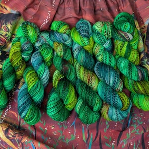 May include: Several skeins of hand-dyed yarn in shades of green, yellow-green, and teal. The yarn is arranged in a row, showcasing the color variations. The background features a fabric with a creature design and a red floral pattern.