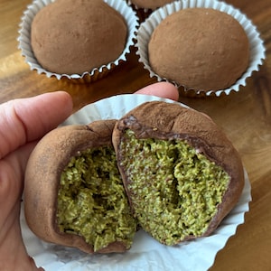 May include: Close-up of chocolate truffles. The truffles are round, coated in cocoa powder, and sit in white paper cups. One truffle is cut in half, revealing a green filling. The truffles are on a wooden surface.