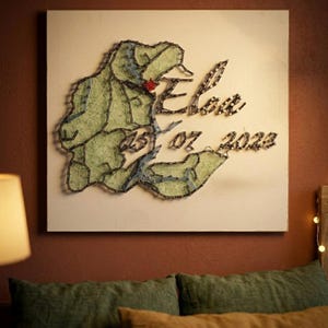 May include: A handcrafted wall hanging with a map design made from green and blue thread on a white canvas. The word "Elan" and the date "25 07 2023" are spelled out in thread. A small red detail is present.