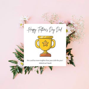 Può includere: Un biglietto per la festa del papà con un'illustrazione di un trofeo d'oro e la scritta "Happy Father's Day Dad". Il biglietto include anche la scritta "this card has more trophies than your club this year! (which isn't hard)". Il biglietto è circondato da fiori rosa e bianchi.