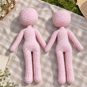 May include: Two light pink crochet dolls with round heads, slender arms, and legs. The dolls are crafted with a visible yarn stitch pattern, featuring a simple, featureless design without facial details or clothing, positioned side by side.