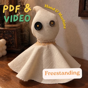 May include: A beige fabric ghost doll with two mismatched black button eyes, yellow cross-stitch detailing at the neck, and a flared base. Text on the image reads PDF & VIDEO, Hand & Machine, and Freestanding. The doll is made of light-coloured linen.