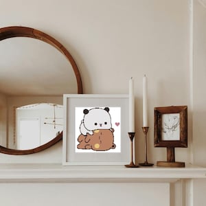 Op de afbeelding: Een wit ingelijst print met een cartoonillustratie van een pandabeer die een bruine beer omhelst. De panda heeft een hartvormige neus en de beren glimlachen. De print is op een witte achtergrond en heeft een roze hart in de rechterbovenhoek.