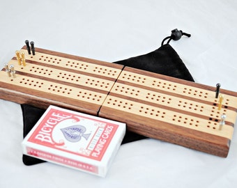 Puede incluir: Un tablero de cribbage de madera con acabado de nogal y arce. El tablero está doblado por la mitad y viene con una bolsa de cordón negro. También se incluye una baraja de cartas Bicycle.