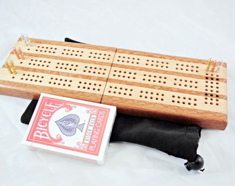 Könnte beinhalten: Ein hölzernes Cribbage-Brett mit einer hellen Holzoptik und dunklen Holzstreifen. Das Brett ist geöffnet und hat Stifte in den Löchern. Ein Deck Bicycle-Spielkarten liegt neben dem Brett.