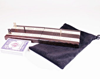 Könnte beinhalten: Ein hölzernes Cribbage-Spielbrett mit dunkelbraunem und weiß gestreiftem Design, Metallstiften und einem Deck Bicycle-Spielkarten. Das Brett ist gefaltet und wird von einem schwarzen Samtbeutel begleitet.