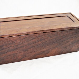 May include: A rectangular wooden box with a dark brown finish. The box has a lid with a recessed panel and a simple, elegant design. The wood grain is visible, adding to the natural aesthetic.