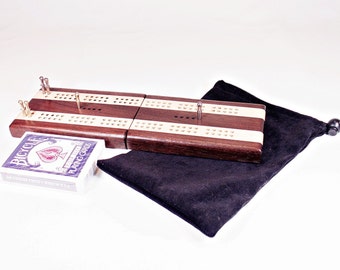 Könnte beinhalten: Ein Cribbage-Brett aus Holz mit Walnuss- und Ahornfurnier. Das Brett ist geschlossen und wird mit einer schwarzen Samt-Tragetasche und einem Kartenspiel geliefert.