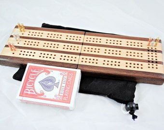 Könnte beinhalten: Ein Cribbage-Brett aus Holz mit Walnuss- und Ahornfurnier. Das Brett ist geschlossen und hat eine schwarze Tragetasche und ein Deck Bicycle-Spielkarten daneben.
