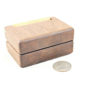 Può includere: Una piccola scatola di legno con un coperchio incernierato. La scatola è in legno scuro e ha un intarsio in legno chiaro sulla parte superiore. La scatola è chiusa e si trova su una superficie bianca.
