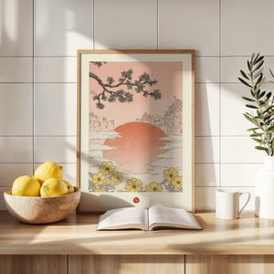 Puede incluir: Una impresi&oacute;n enmarcada de una ilustraci&oacute;n de estilo japon&eacute;s que representa una puesta de sol sobre una masa de agua con una rama de &aacute;rbol con hojas en la esquina superior izquierda. La impresi&oacute;n est&aacute; rodeada de un marco de madera claro y se encuentra sobre una superficie de madera.